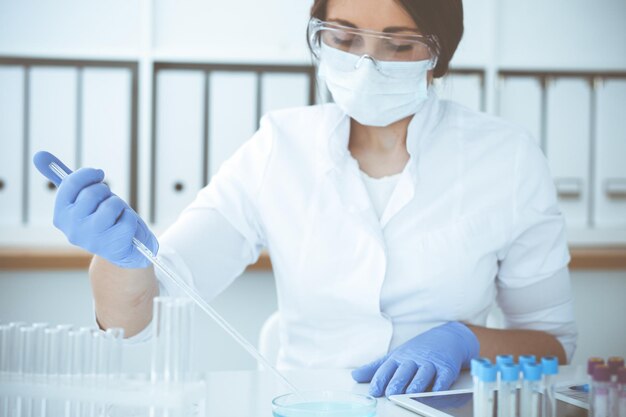 Крупный план профессиональной женщины-ученого в защитных очках, проводящей эксперимент с реагентами или анализом крови в лаборатории. Концепция медицины, биотехнологии и исследований.