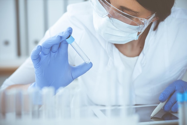 Close-up of professional female scientist in protective eyeglasses making experiment with reagents or blood test in laboratory. Medicine, biotechnology and research concept.