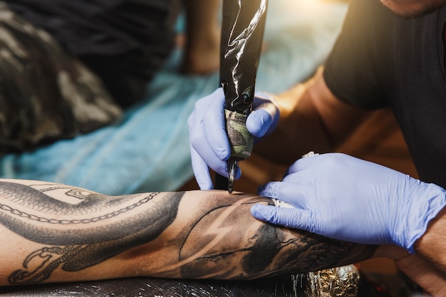 Close up of professional tattooer artist doing tattoo on the arm of a young man by machine with black ink