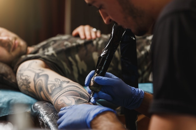 Close up of professional tattooer artist doing tattoo on the arm of a young man by machine with black ink