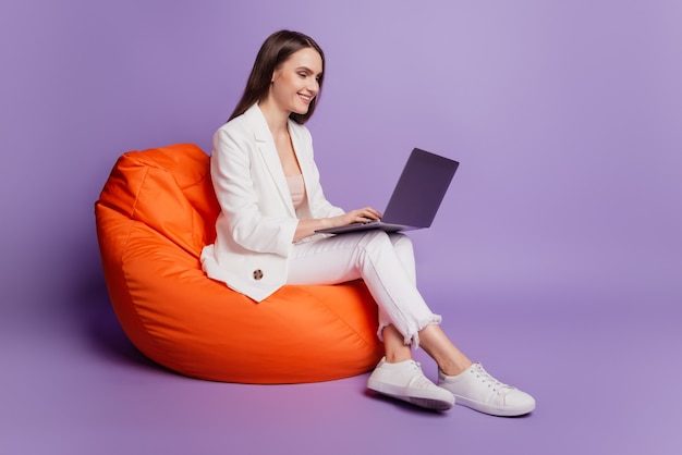 Close up profile portrait of positive lady sit beanbag hold computer typing on purple wall