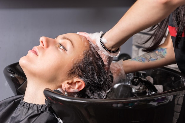 Photo close up profile view of young brunette woman having hair washed and soaped by stylist in washing sink in beauty salon