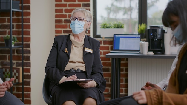 Close up of psychologist with face mask talking to patients at aa therapy meeting. Woman therapist leping people with alcohol addiction and mental health problems during pandemic.