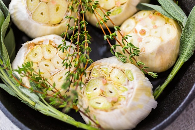 Close up of raw garlic heads with fresh spices in cast iron skillet for preparing roasted garlic recipe.