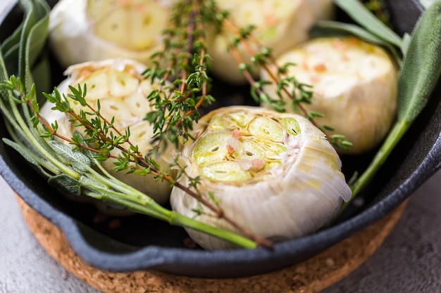 Close up of raw garlic heads with fresh spices in cast iron skillet for preparing roasted garlic recipe.