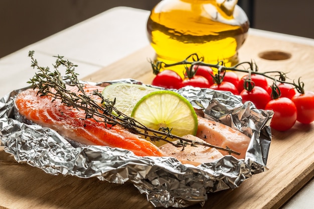 Close up of the raw red fish slice with thyme and lime lying in foil and branch of cherry tomatoes on the cutting board near