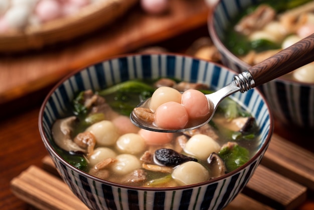 Close up of red and white tangyuan (tang yuan, glutinous rice dumpling balls) with savory soup in a bowl on wooden table background for Winter solstice festival food.