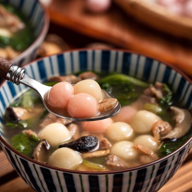 Close up of red and white tangyuan (tang yuan, glutinous rice dumpling balls) with savory soup in a bowl on wooden table background for Winter solstice festival food.