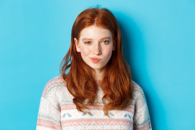 Close-up of redhead cunning girl having an idea, smirk and stare at camera satisfied with plan, standing over blue background