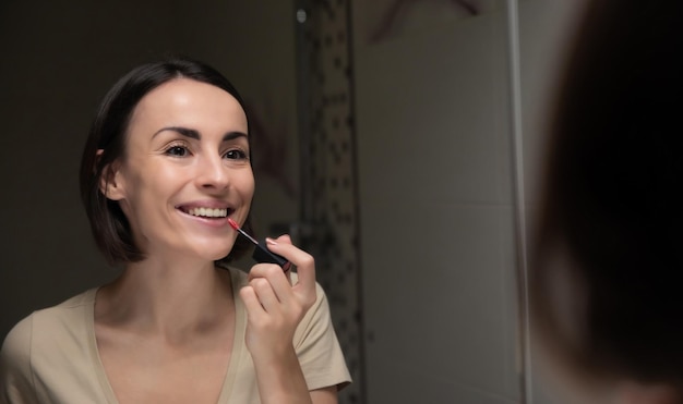Крупный план отражения портрета великолепной улыбающейся брюнетки в зеркале, пока она делает макияж губ в ванной