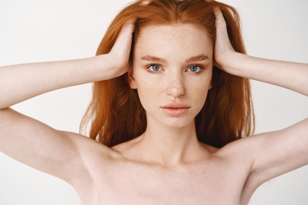 Close-up of relaxed young redhead woman with pale skin and freckles, massaging natural red hair, standing naked without make-up on white wall