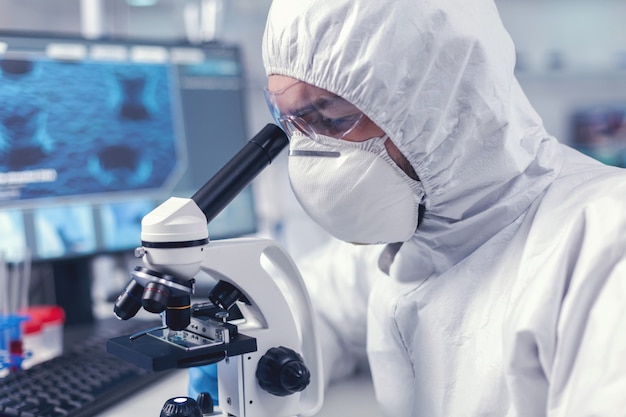 Close up of researcher looking throug microscope in biochemistry laboratory. Virolog in coverall during coronavirus outbreak conducting healthcare scientific analysis.