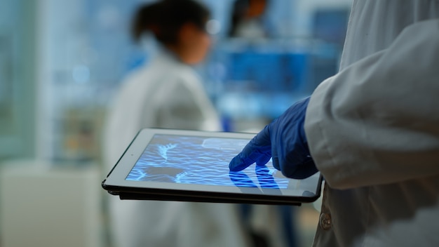 Close up of researcher using tablet, pointing, analysing DNA informations standing in medical laboratory. Viorolog doctor working to discover medical treatment, vaccine development, virus evolution