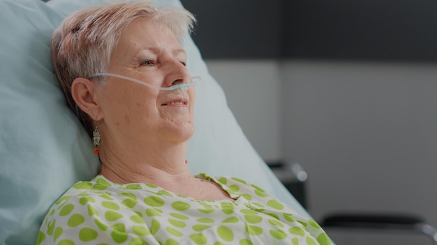 Photo close up of retired patient with illness having nasal oxygen tube and sitting in hospital ward bed. senior woman preparing to be treated against disease, hoping for recovery at clinic.