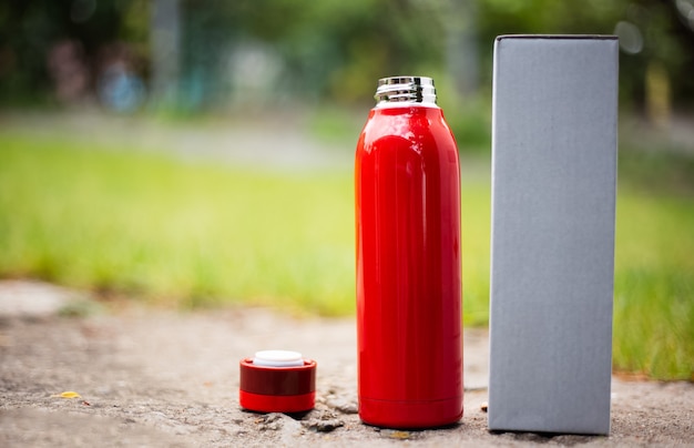 Primo piano della bottiglia termica rossa d'acciaio riutilizzabile per l'acqua accanto al tappo e alla scatola di cartone per l'imballaggio. sfondo sfocato all'aperto.