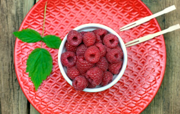  Close-up ripe raspberries in a bowl on a red ceramic plate.