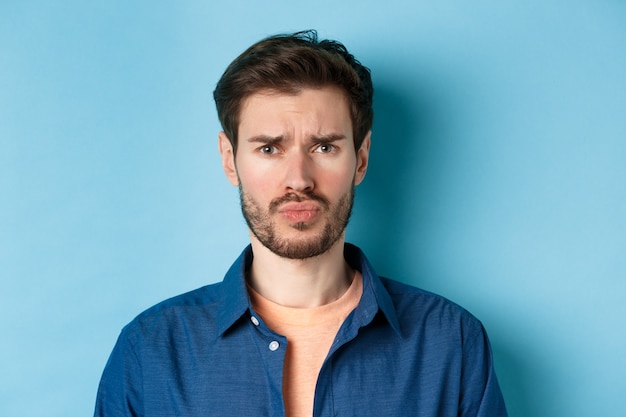 Close up of sad and offended young man sulking at someone, frowning upset, standing on blue background.