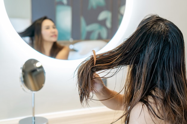 Close up of sad young woman using wooden comb brush her damaged and tangled wet hair in front of mirror