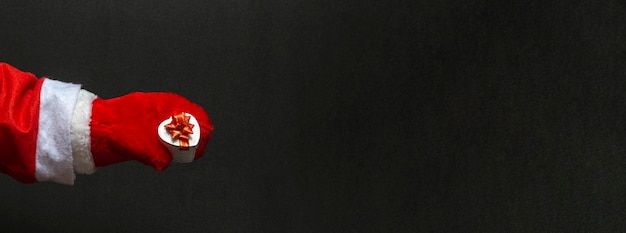 Primo piano di babbo natale in guanti rossi che tiene un regalo su uno sfondo nero con spazio di copia. banner
