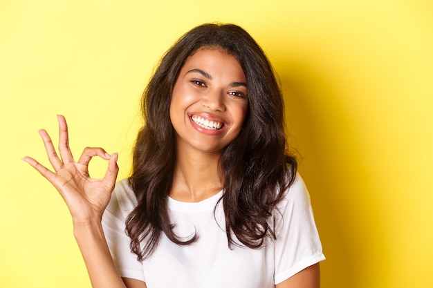 Close-up of satisfied african-american woman, showing okay sign and smiling, recommending something good, standing over yellow background.