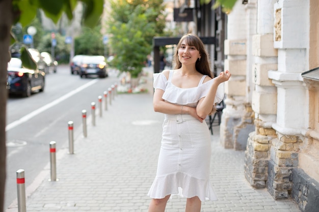 여름이면 거리와 도로를 배경으로 하얀 드레스를 입고 갈색 긴 머리를 한 만족스러운 소녀의 클로즈업