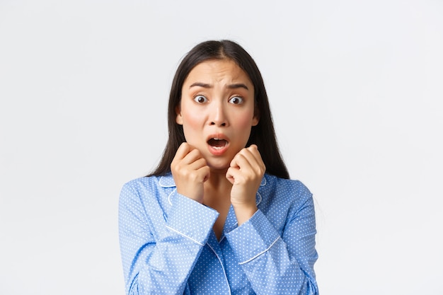 Close-up of scared asian woman in panic standing in pajama speechless, react to frightening and shocking scene, looking afraid, shivering from fear over white background.
