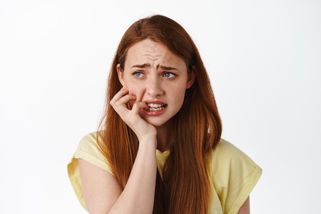Close up of scared and insecure redhead teenage girl biting fingers and looking left at logo in panic standing worried and alarmed nervous of consequences white background