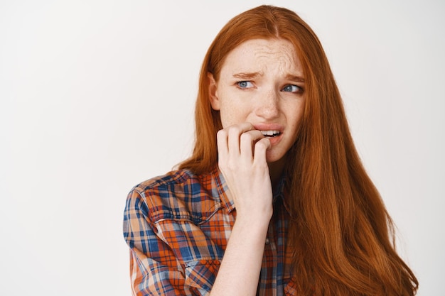 Close-up of scared and nervous woman with red hair, biting fingernails and frowning insecure, looking left worried, standing over white wall