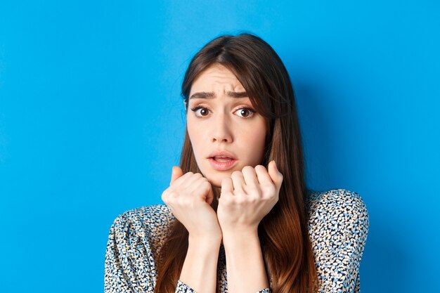 Close-up of scared timid girl looking afraid, shaking from fear, press hands to body and staring frightened, standing on blue.