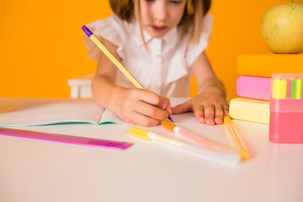 Close-up schoolgirl in a white blouse is sitting at a table and writing in a notebook on a yellow background with a copy of the space