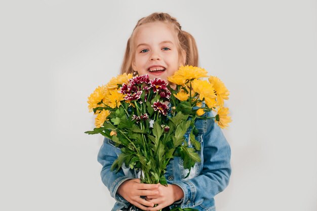 Close-up, selectieve aandacht. Klein blond meisje in een houdt chrysanten en gerbera's in haar handen op een witte achtergrond, een kindmeisje glimlacht en houdt lentebloemen in haar handen