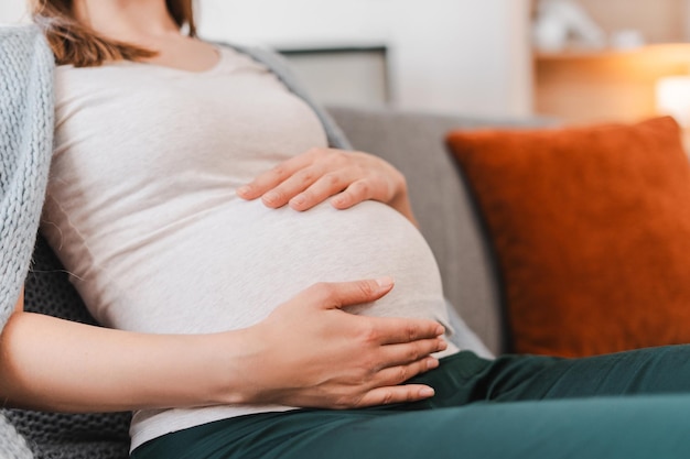 Close up selective focus on belly of pregnant woman touching hands sitting on sofa in cozy home