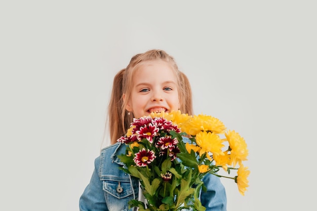 Close up, selective focus. Little blonde girl in an holds chrysanthemums and gerberas in her hands on a white background, a child girl smiles and holds spring flowers in her hands