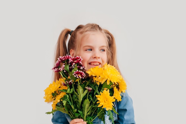 Close up, selective focus. Little blonde girl in an holds chrysanthemums and gerberas in her hands on a white background, a child girl smiles and holds spring flowers in her hands