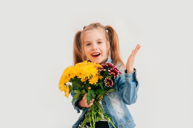 Close up, selective focus. Little blonde girl in an holds chrysanthemums and gerberas in her hands on a white background, a child girl smiles and holds spring flowers in her hands