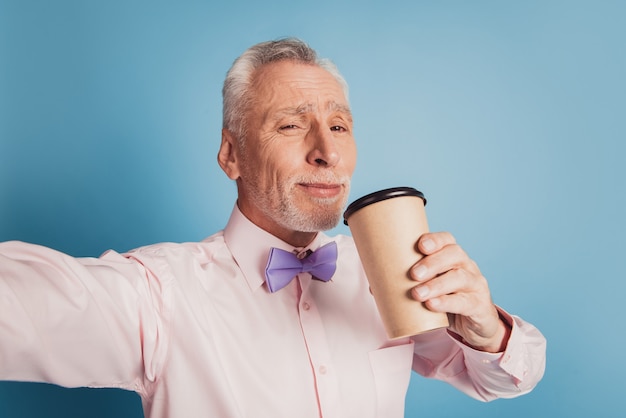 Close up self portrait of senior man drinking takeout coffee on blue backdrop