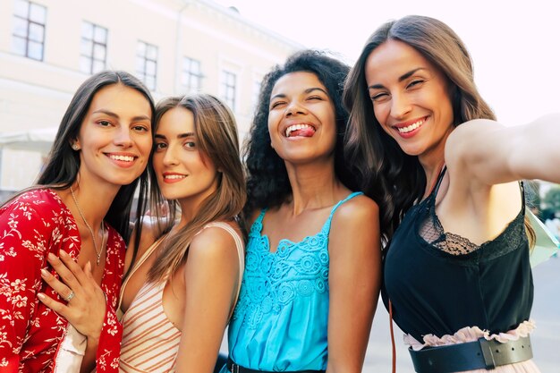 Close-up selfie foto van vier vriendinnen op vakantie, zomeroutfits dragen, kijken naar de camera, breed glimlachen en de tong uitsteken.