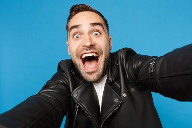 Close up selfie of stylish young unshaven man in black leather jacket white t-shirt looking camera isolated on blue wall background studio portrait. People sincere emotions concept. Mock up copy space