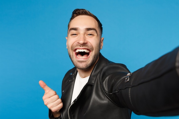 Close-up selfie van stijlvolle jonge ongeschoren man in zwart lederen jas wit t-shirt op zoek camera geïsoleerd op blauwe muur achtergrond studio portret. Mensen oprecht emoties concept. Bespotten kopie ruimte