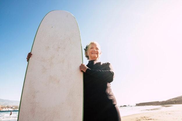 Close up of senior at the beach with wetsuit and surftable ready to go to try her first class of surf - mature woman smiling with the sea or the ocean at the background