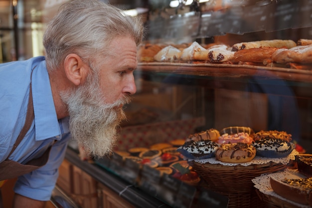 Close up of a senior bearded male baker looking at retail\
display at his bakery store