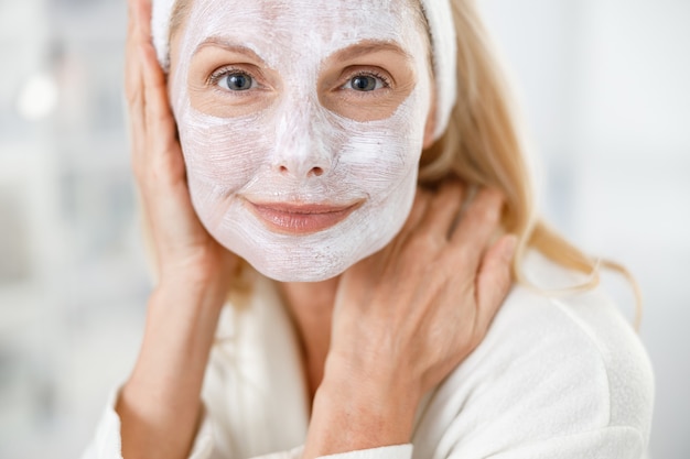 Photo close up of senior blond woman with nutrition mask on her face, visiting cosmetologists in modern medical center.