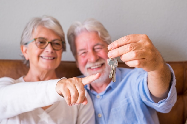 Закройте старшие пары показывая ключи от дома. Пожилая пара держит ключи для инвестиций в недвижимость. Довольная старая пара держит ключи от своего нового дома
