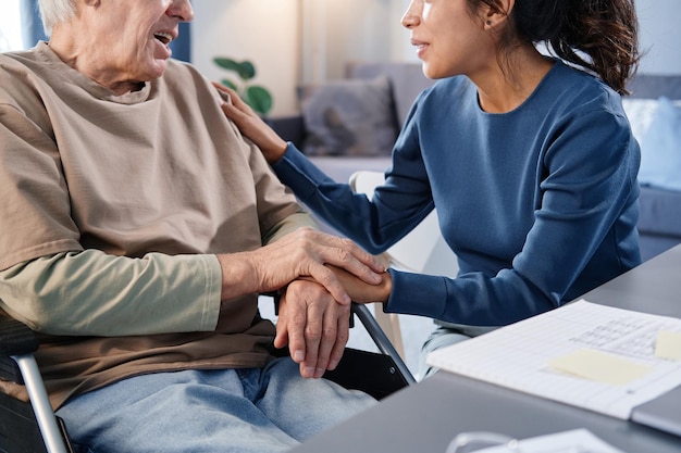 Photo close-up of senior man using wheelchair talking to the nurse while she holding his hands at the table