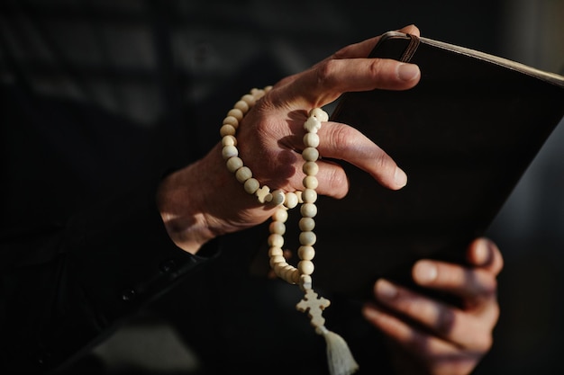 Foto primo piano del sacerdote anziano che tiene la bibbia e il rosario che pregano alla luce della drammaticità