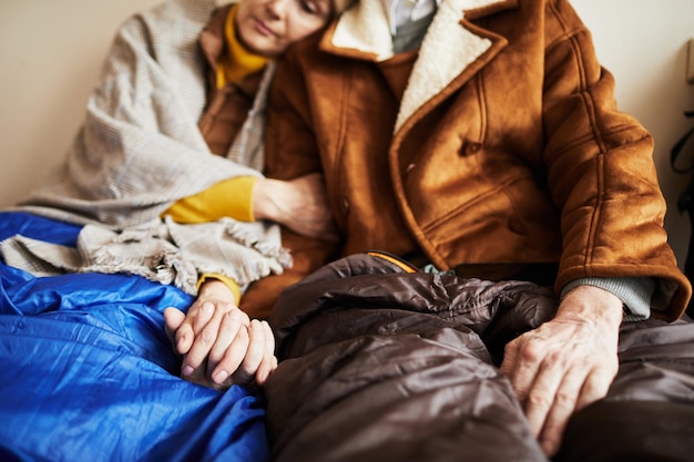 Close up of senior refugee couple hiding in shelter and holding hands in hope during war or crisis c