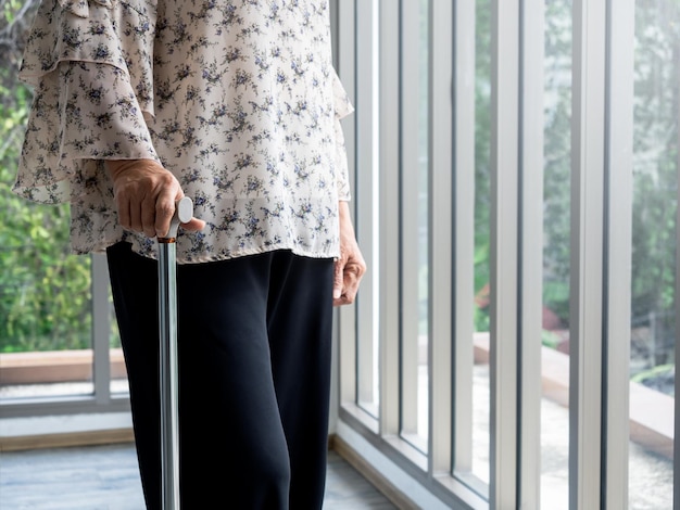 Primo piano della mano della donna anziana che tiene un bastone da passeggio in piedi da solo vicino alla finestra di vetro nella stanza con spazio per la copia donna anziana sola la nonna con un bastone da passeggio