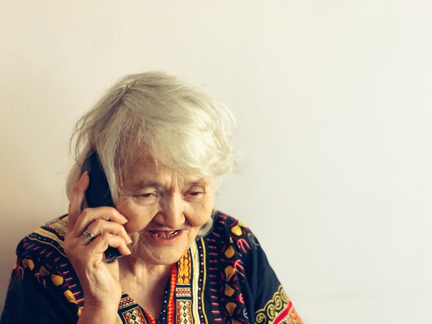 Photo close-up of senior woman talking on phone by white wall