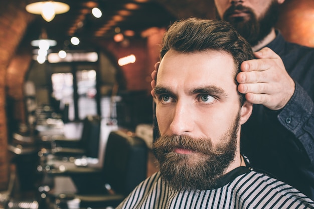 Close up of serious and bearded guy sitting in chair and looking straight forward. His hairdresser is modelling his hair.