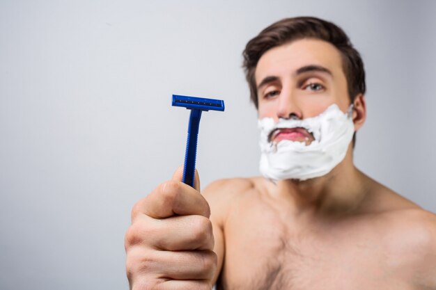 Close up of serious and concentrated guy that stands in white room and showing a good razor that he has. He has put some foam on his beard and the man is ready to get sheved. Cut view.